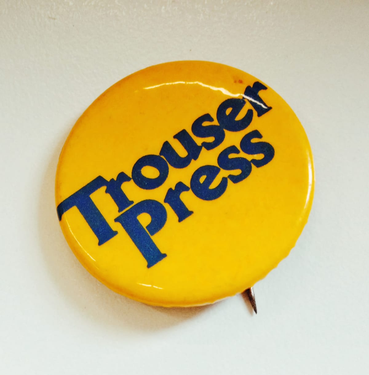 A yellow pin-back button lying on a pale grey surface, the words "Trouser Press" printed across it in heavy navy lettering on two stacked lines set at a diagonal, the pin's clasp just visible beneath the lower edge.