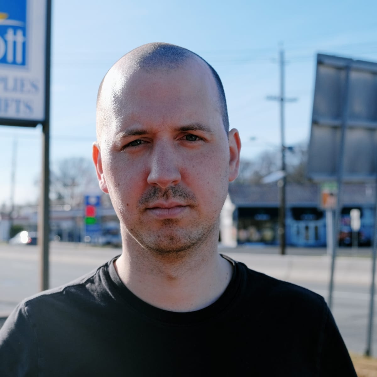 Jason Calhoun looks directly at the camera in a black t-shirt, close-cropped hair, a roadside sign and storefronts blurred behind him under a clear sky.