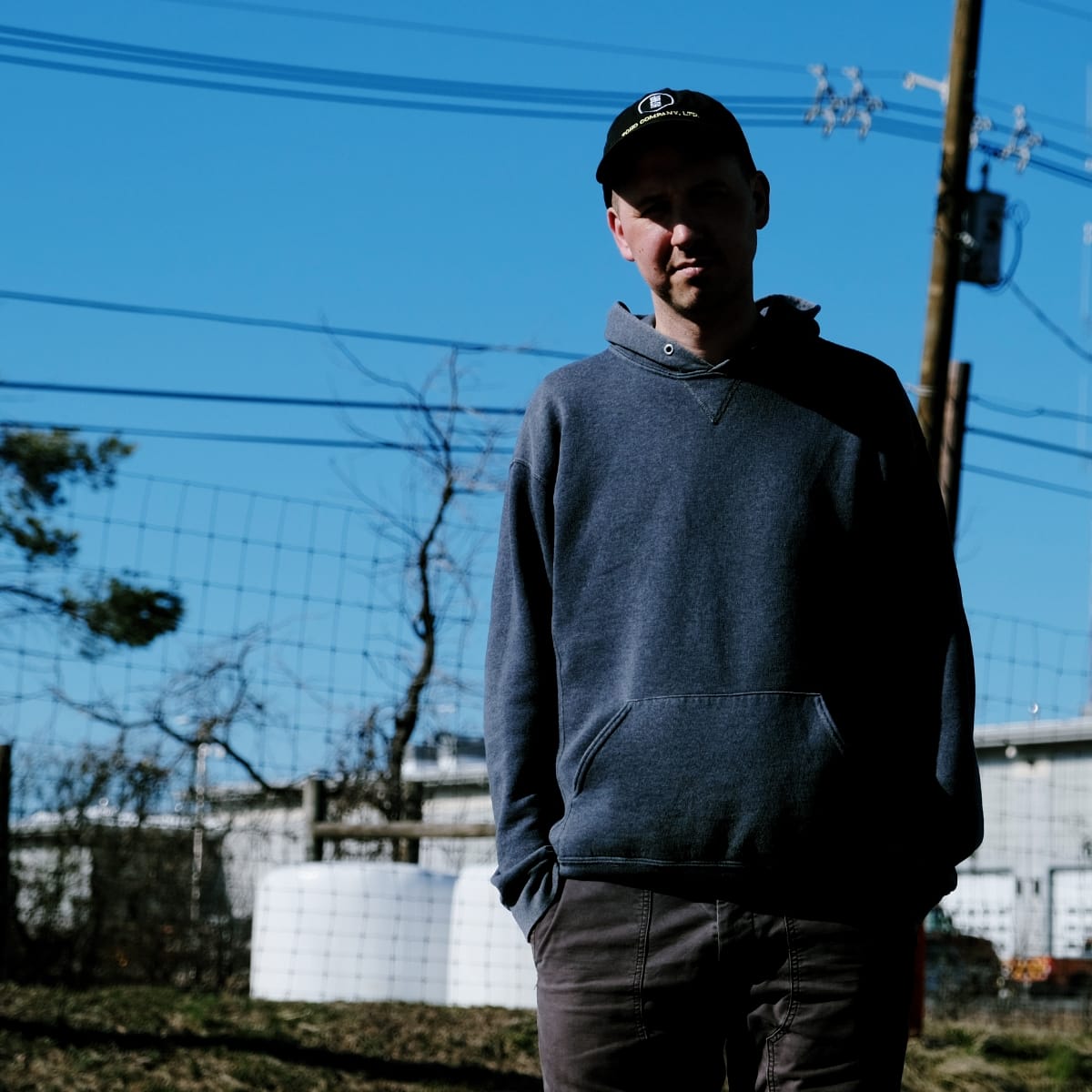 Jason Calhoun stands under power lines against a bright blue sky, hands in the pockets of a grey hoodie and wearing a dark cap, a wire fence and white storage tanks behind him.