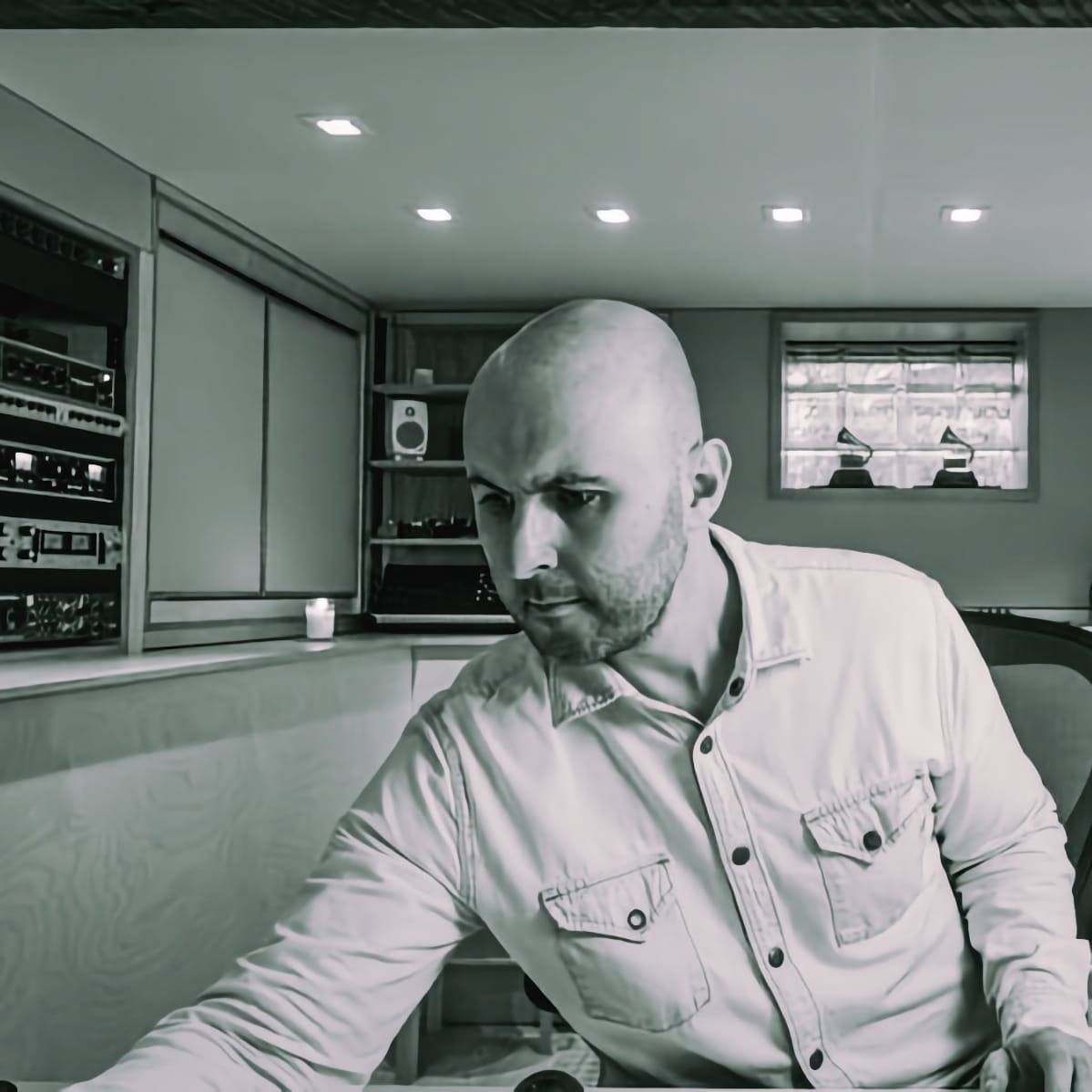 Joseph Branciforte looks down at a console in greyfade Studio, rack gear and a monitor speaker behind him, two Grammy statuettes on a windowsill to his right. Black and white.
