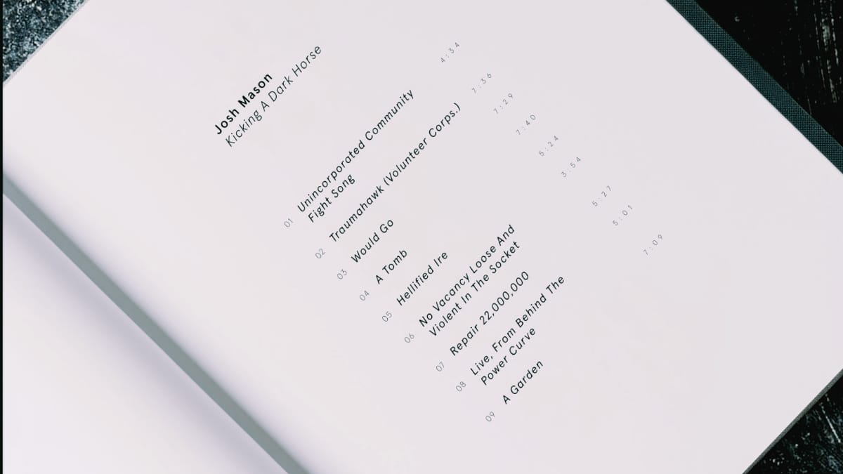 A pale book page photographed at an angle, its rotated type listing Josh Mason's Kicking A Dark Horse and all nine track titles with running times.