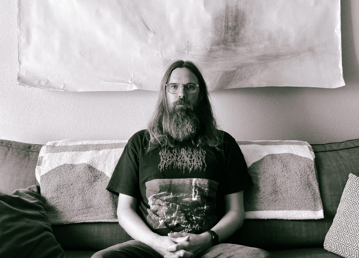 Josh Mason, long-haired and heavily bearded, sits on a sofa with hands clasped, wearing glasses and a dark band T-shirt, a large unframed paper work on the wall above. Black and white.