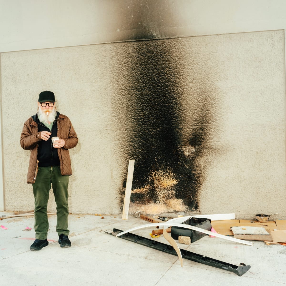 Kurt Wagner stands against a pale stucco wall marked by a large black scorch stain, lighting a cigarette and holding a paper cup. Burned debris lies scattered on the concrete beside him.