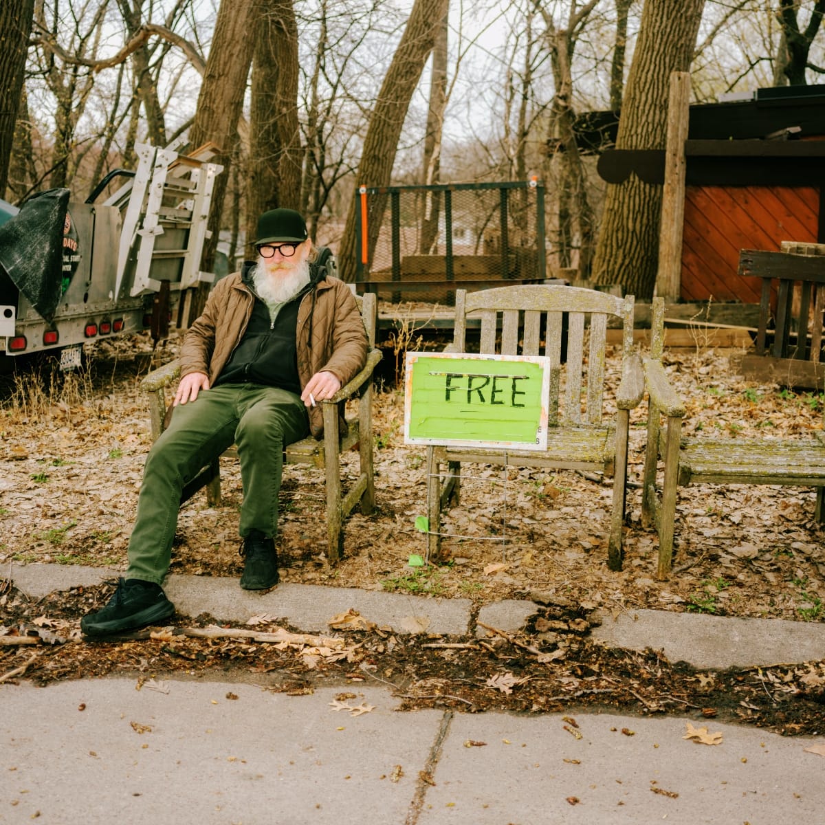 Kurt Wagner sits on a weathered wooden bench among bare trees and dead leaves, a cigarette in one hand. A hand-painted green sign reading FREE leans against the bench beside him.