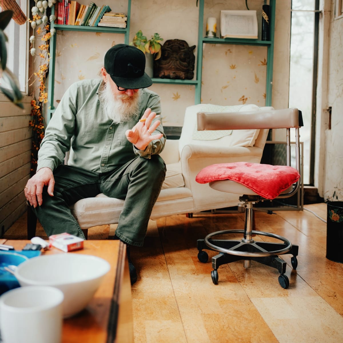 Kurt Wagner leans forward on a white sofa, cigarette raised in one hand, black cap low over his glasses and long white beard. A red-cushioned rolling stool sits on the floor beside him.