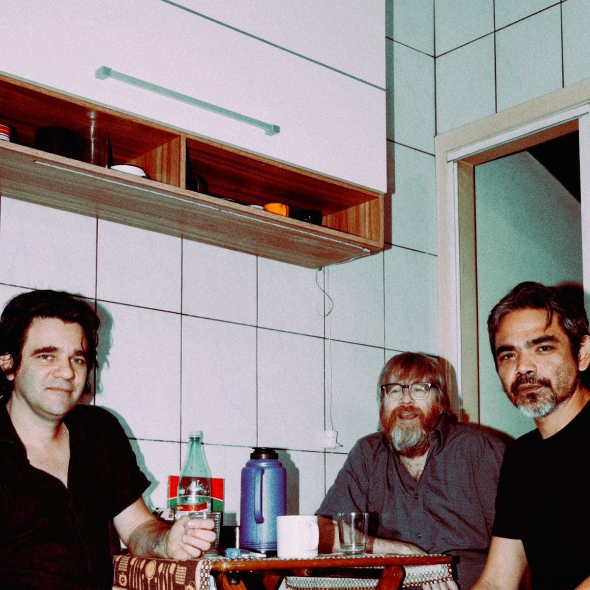 Riviera Gaz around a small table in a green-tiled kitchen — Gustavo Riviera at left with a glass, Steve Shelley centre, Paulo Kishimoto at right, a bottle and blue thermos between them.