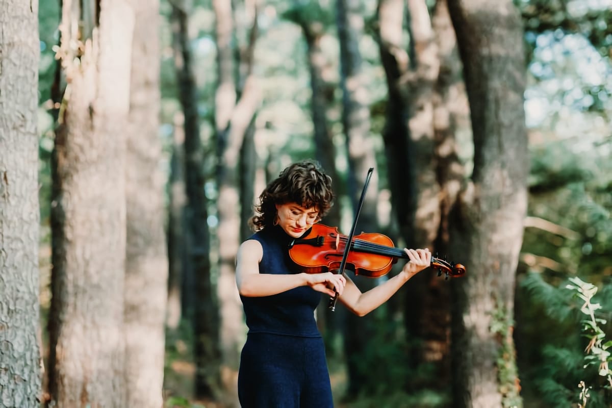 Sophie Wellington plays fiddle among tall trees in a forest, looking down at her hands, dressed in a sleeveless navy top.
