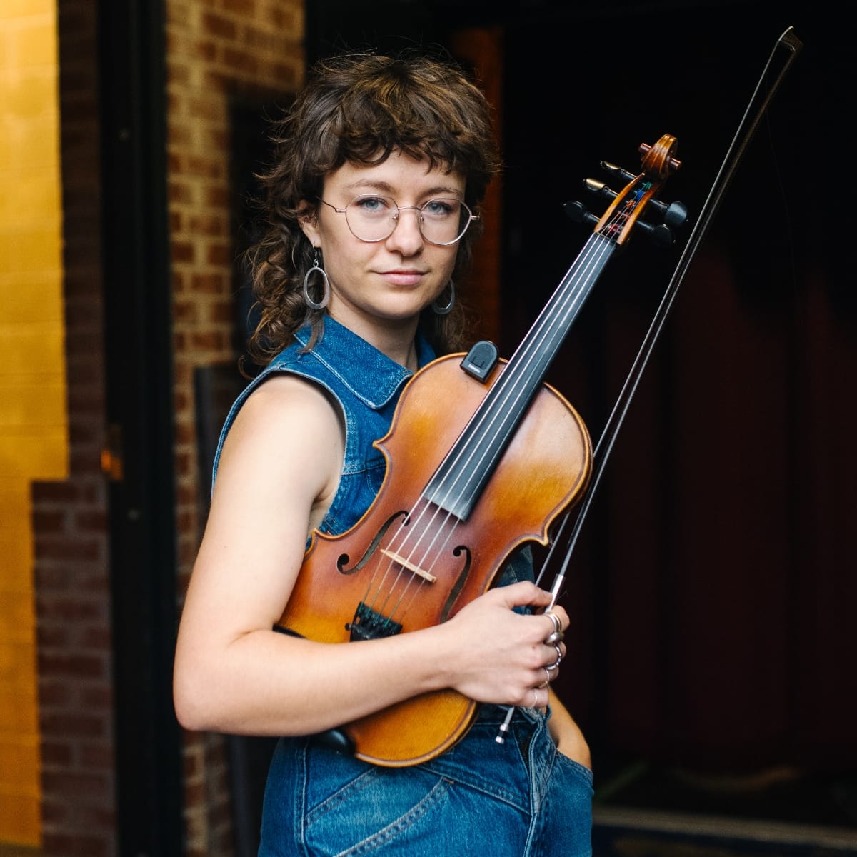 Sophie Wellington stands in a doorway holding a fiddle and bow against her chest, in a sleeveless denim outfit and round glasses, with brick and a yellow wall behind her.