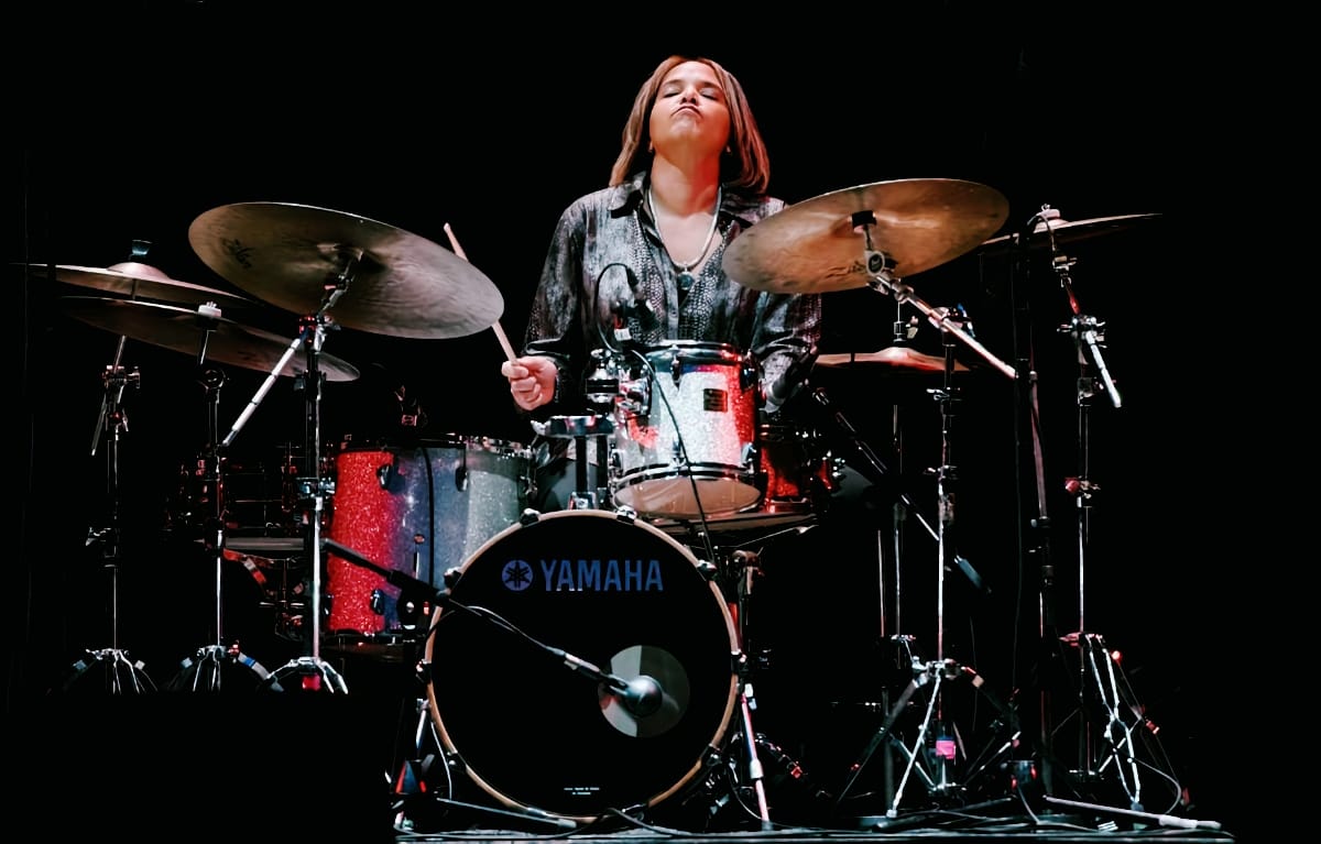 Terri Lyne Carrington plays a sparkle-finish Yamaha kit on a darkened stage, eyes closed and stick raised, cymbals and stands framing her against a black backdrop.