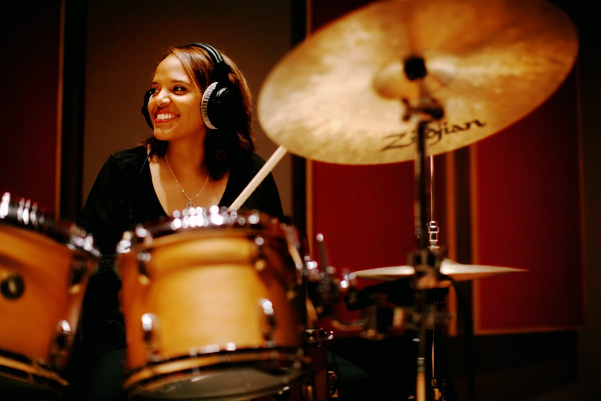 Terri Lyne Carrington sits behind a drum kit in a studio, wearing headphones and smiling off to one side, a large ride cymbal filling the right of the frame.
