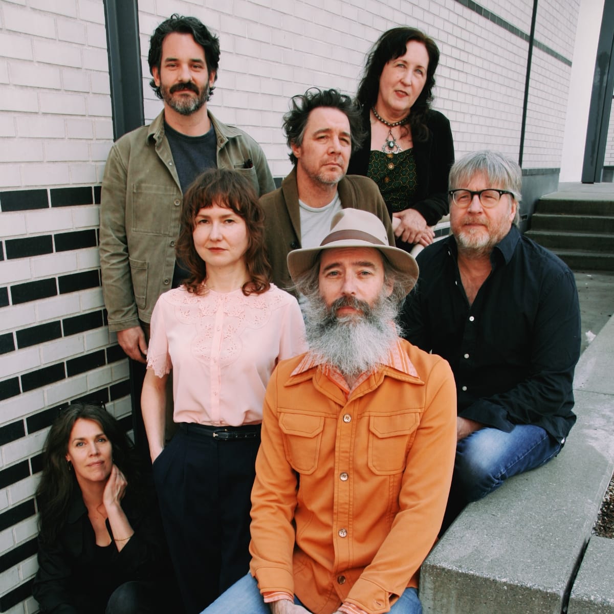 The Odyssey Cult grouped on concrete steps against a black-and-white brick wall, Ethan Miller seated in front in a tan wide-brimmed hat and orange work shirt, long grey beard.