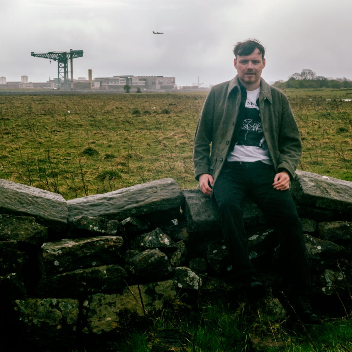 Jamie Livingstone sits on a drystone wall in a rough green field, a shipyard crane and low industrial buildings on the skyline behind him under heavy grey cloud.