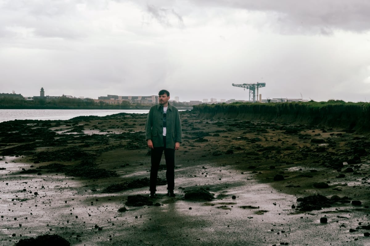 Jamie Livingstone stands alone on a wet, rocky tidal flat, hands at his sides, a shipyard crane and low buildings across the water behind him.