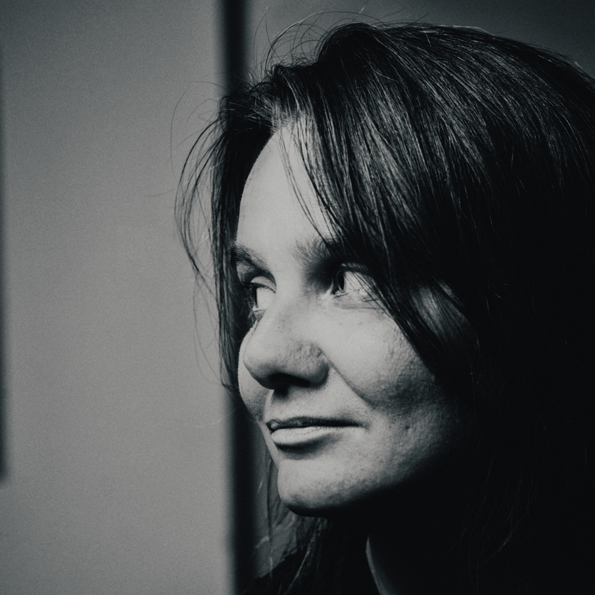 Black-and-white close portrait of Christiane Bopp, turned in profile and looking off to one side with a slight smile, dark hair falling across her face, against a plain grey wall.