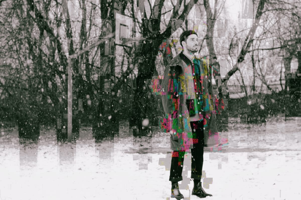 arc rae stands alone in a snow-covered lot beneath bare trees and a basketball hoop, his long coat dissolving into blocks of digital glitch in green, pink, and yellow.