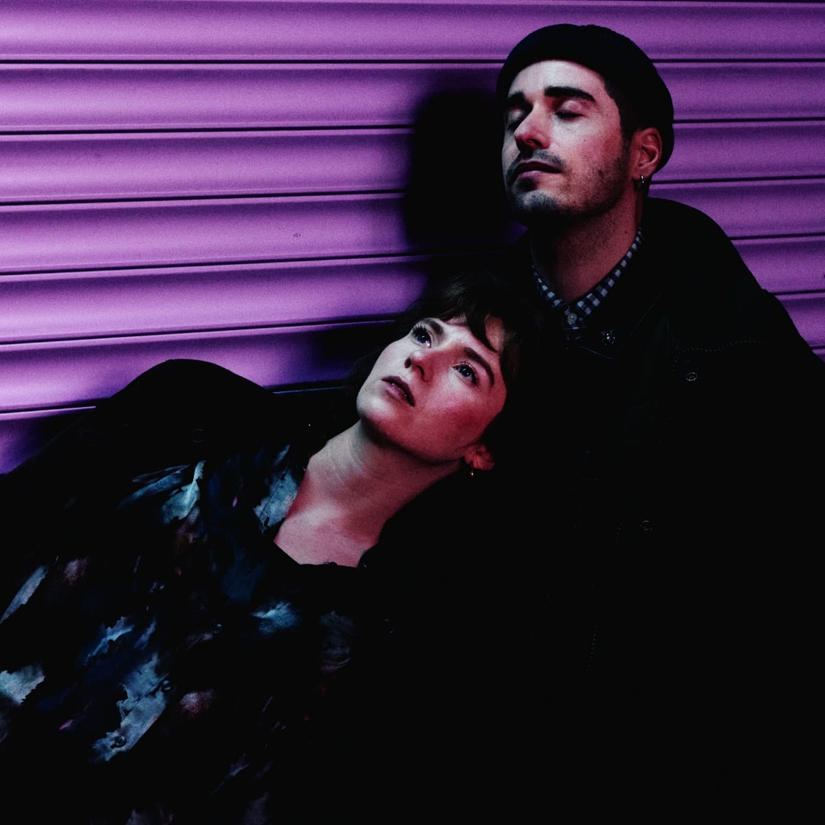 Mathilde Vendramin rests her head against arc rae's shoulder as they sit against a purple corrugated shutter, her eyes open and looking up, his closed.