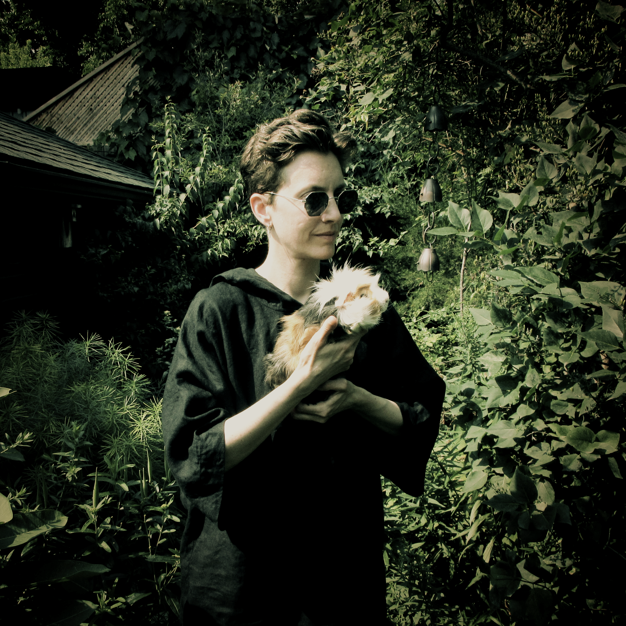 Avi Engel stands in a shaded, overgrown garden wearing dark sunglasses and a black linen hoodie, cradling a long-haired orange-and-white guinea pig against their chest, with wind chimes and a shingled roof behind them.