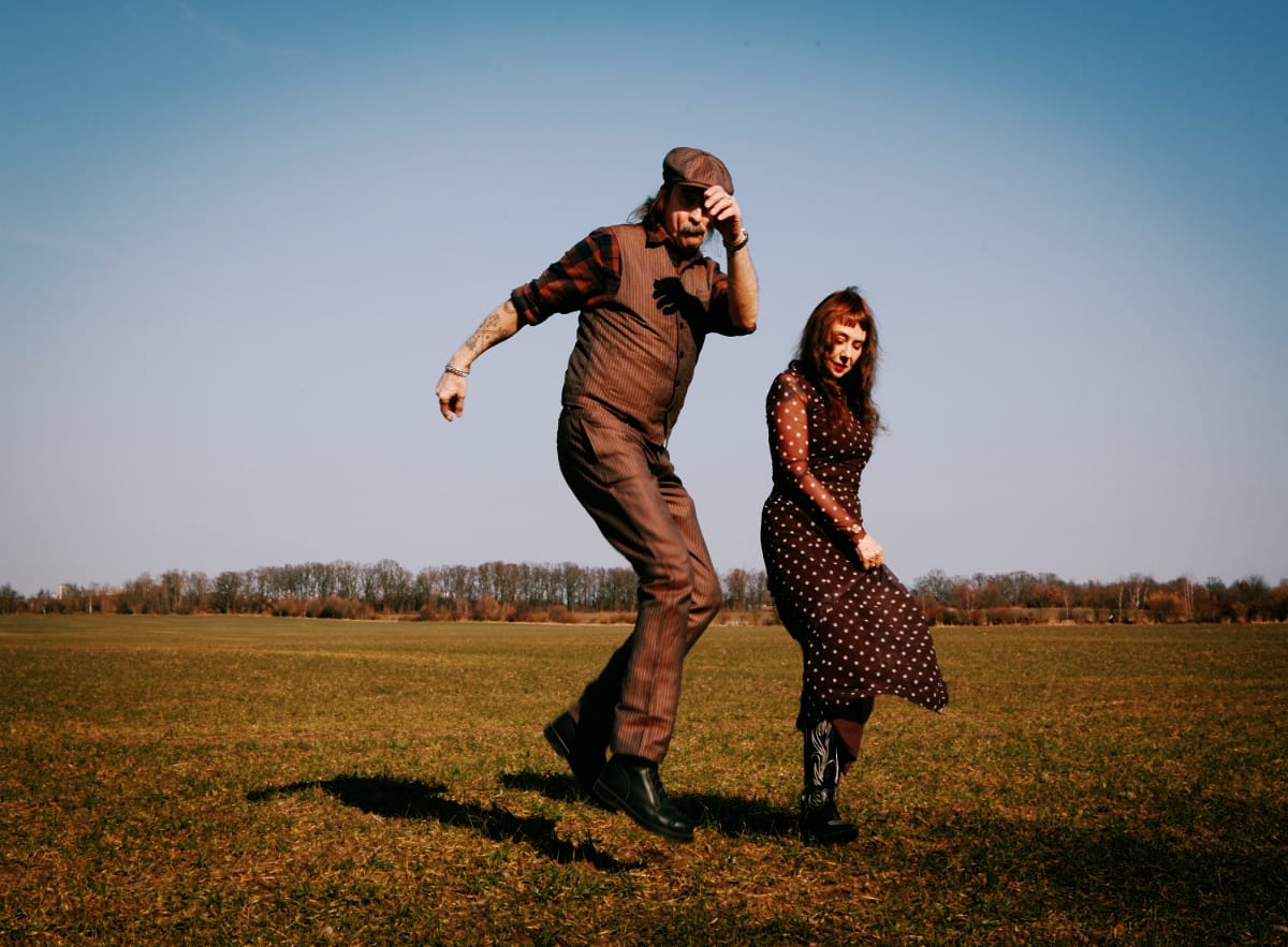 hackedepicciotto dance in a bare field under a clear sky — Alexander Hacke mid-stride with a hand to his cap, Danielle de Picciotto beside him, head down, dress in motion.