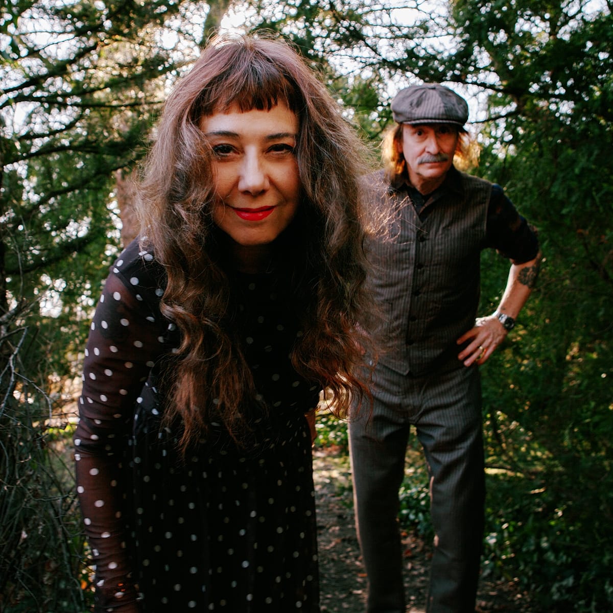 Danielle de Picciotto leans toward the camera on a wooded path, smiling in a polka-dot dress, with Alexander Hacke standing behind her in a flat cap, hands on hips.