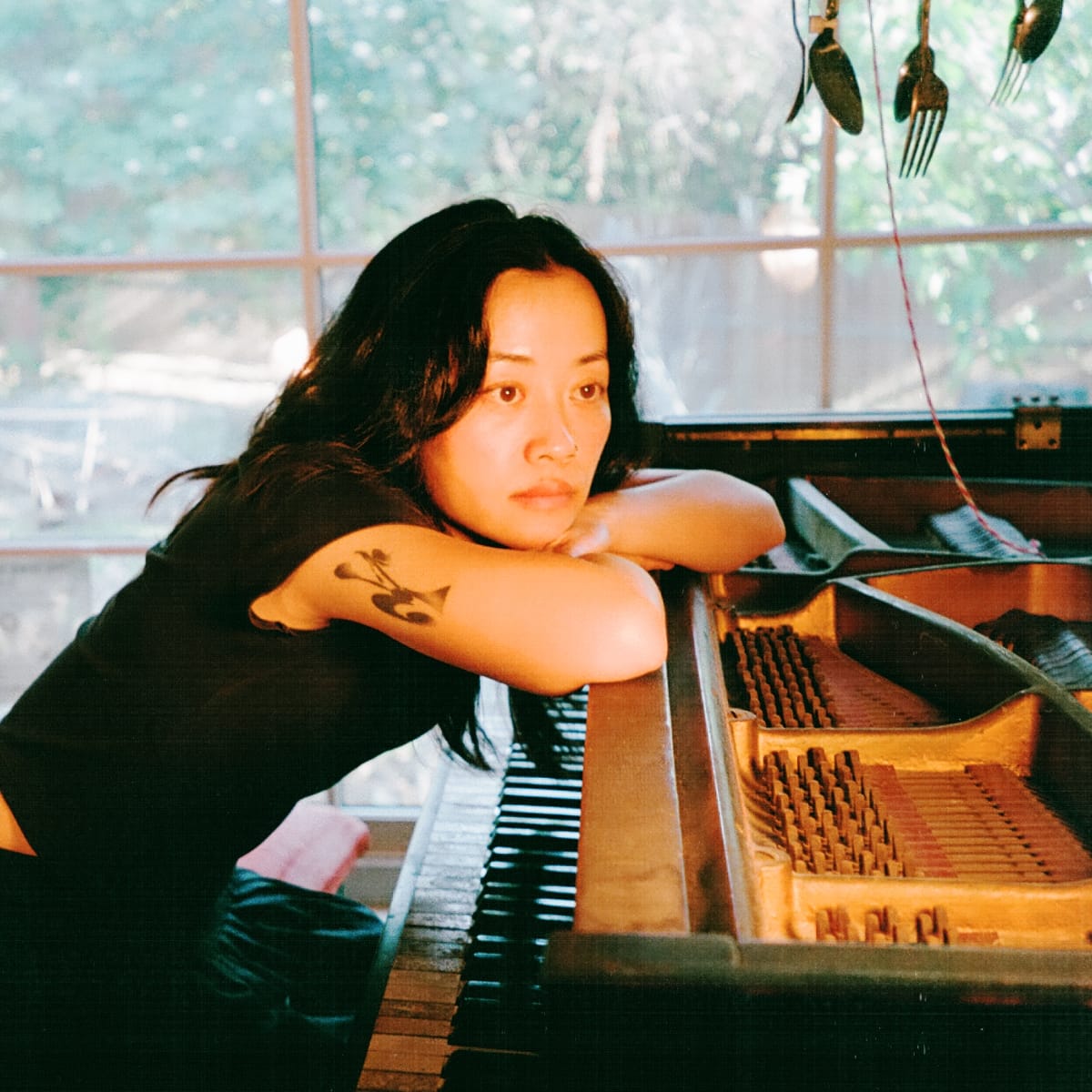 sachi's mirror rests her folded arms on the open case of a piano, hammers and strings exposed, in a sunlit room where mismatched forks and spoons hang on a wire above her.