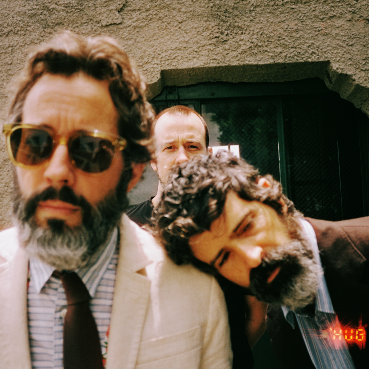 The three members of HUG press together against a stucco wall: one in yellow sunglasses and a pale jacket, one behind in a black tee, the third with his head tilted sideways onto a shoulder.