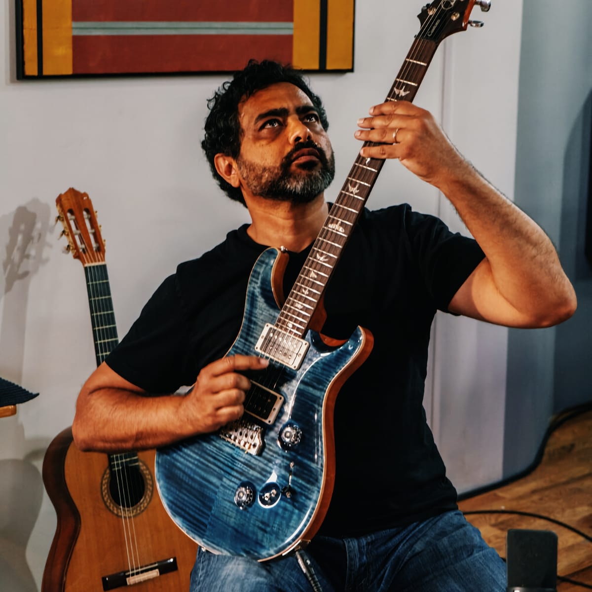 Ayman Fanous sits holding a blue electric guitar upright, gazing up its neck; a nylon-string classical guitar leans behind him beneath a red-and-gold abstract painting.