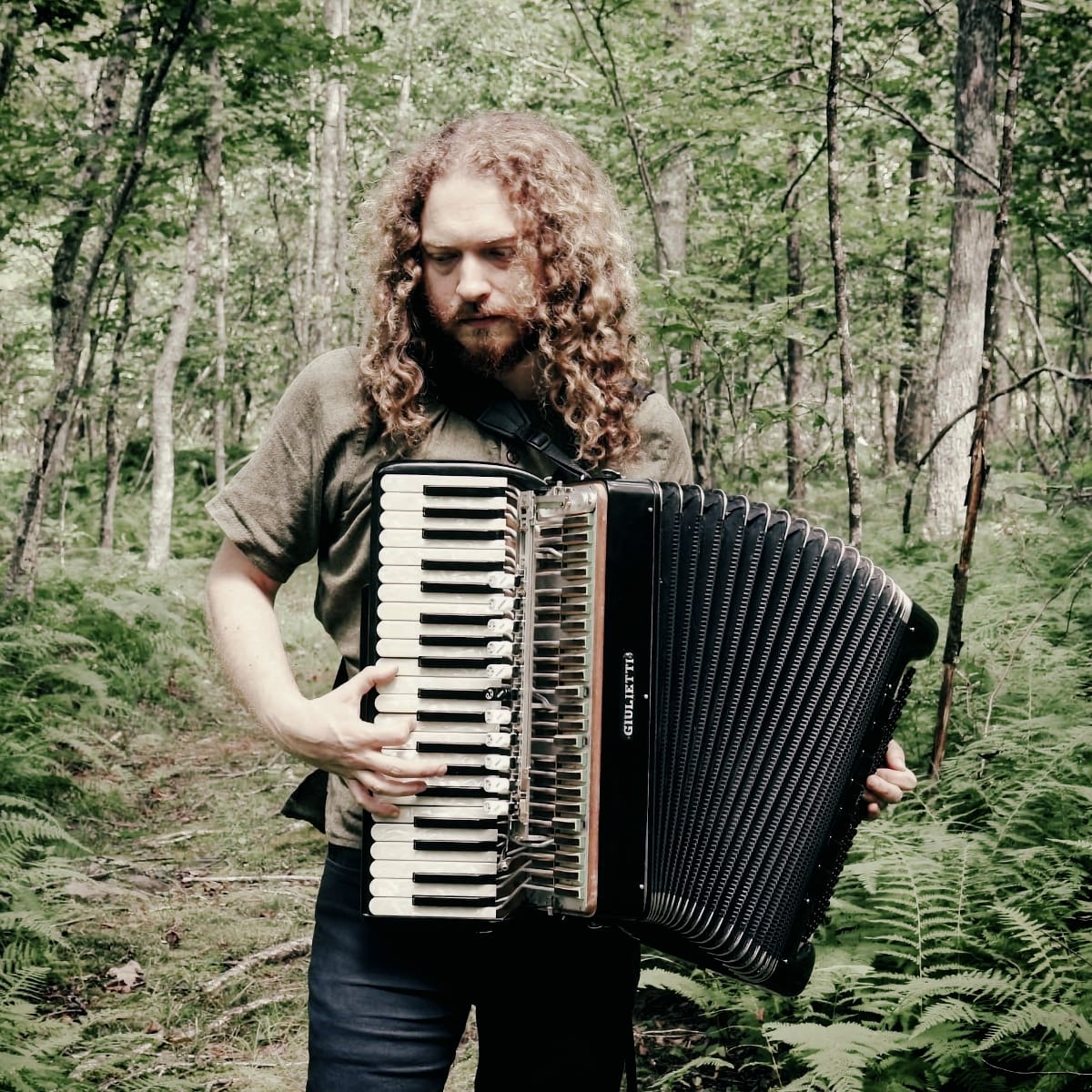 Ben Richter stands among ferns in a green wooded clearing, playing a black Giulietti piano accordion with the bellows half drawn.