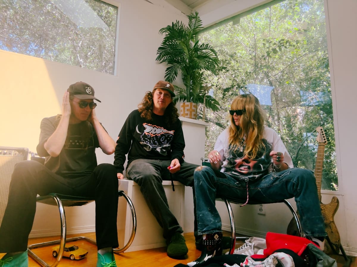 Black Bananas sit in a sunlit room with tall windows and a potted palm: one member cups hands over ears, one talks, and Herrema, in sunglasses and ripped jeans, adjusts her rings.