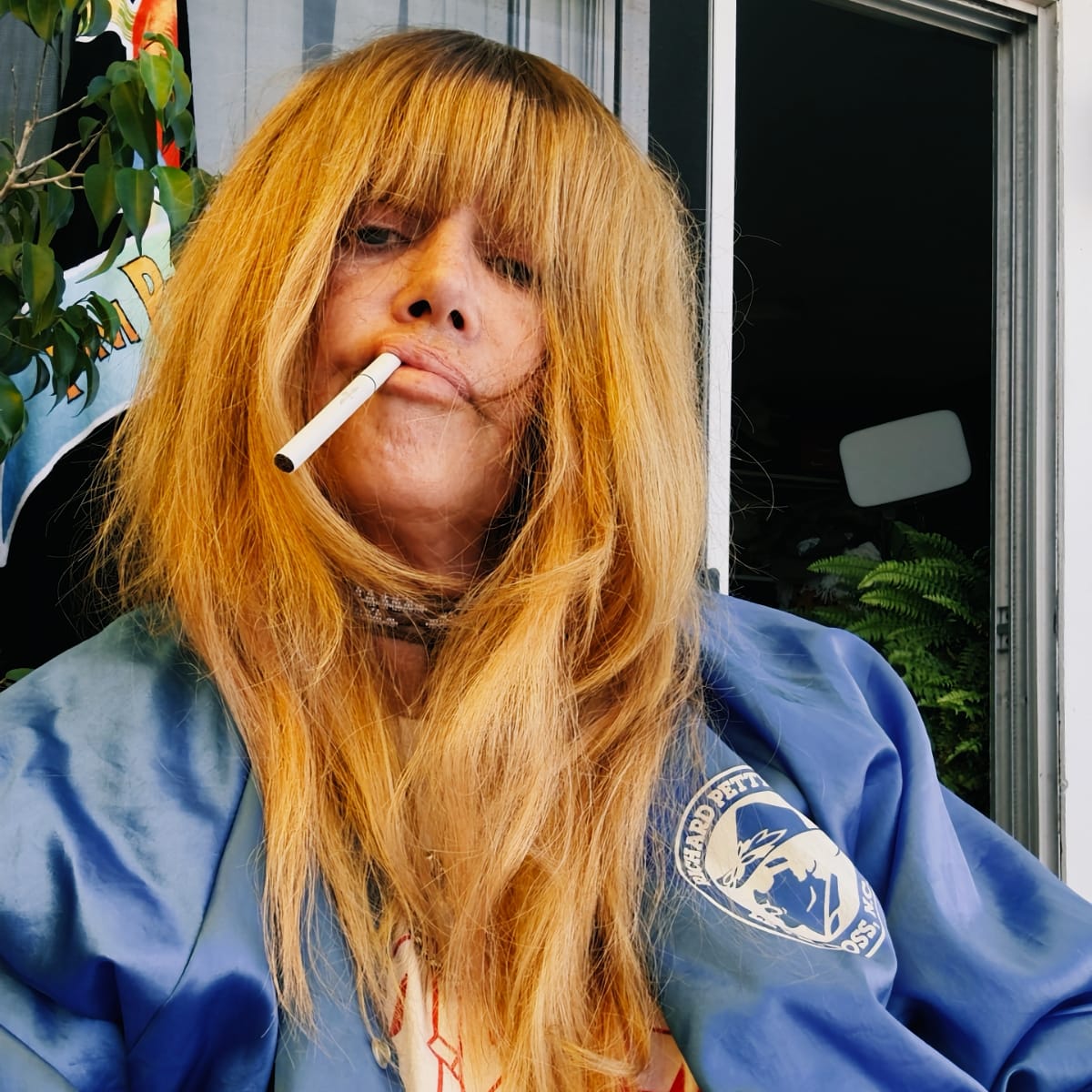 Jennifer Herrema, a cigarette between her lips, looks down at the camera through long strawberry-blond bangs, in a blue satin jacket beside a doorway and potted fern.