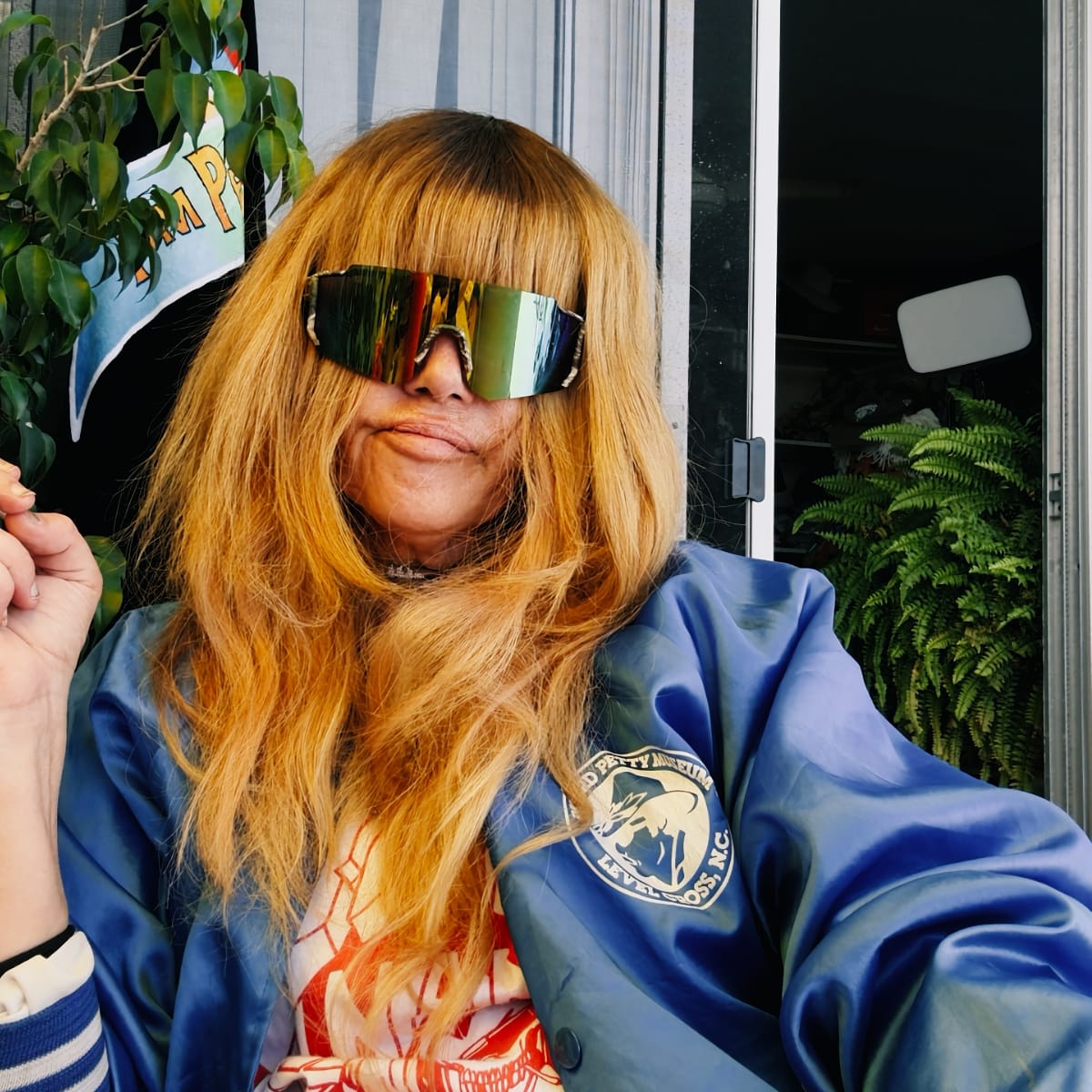 Jennifer Herrema in mirrored shield sunglasses and a blue satin jacket, long bangs falling over the frames, sits beside an open doorway with a fern behind her.