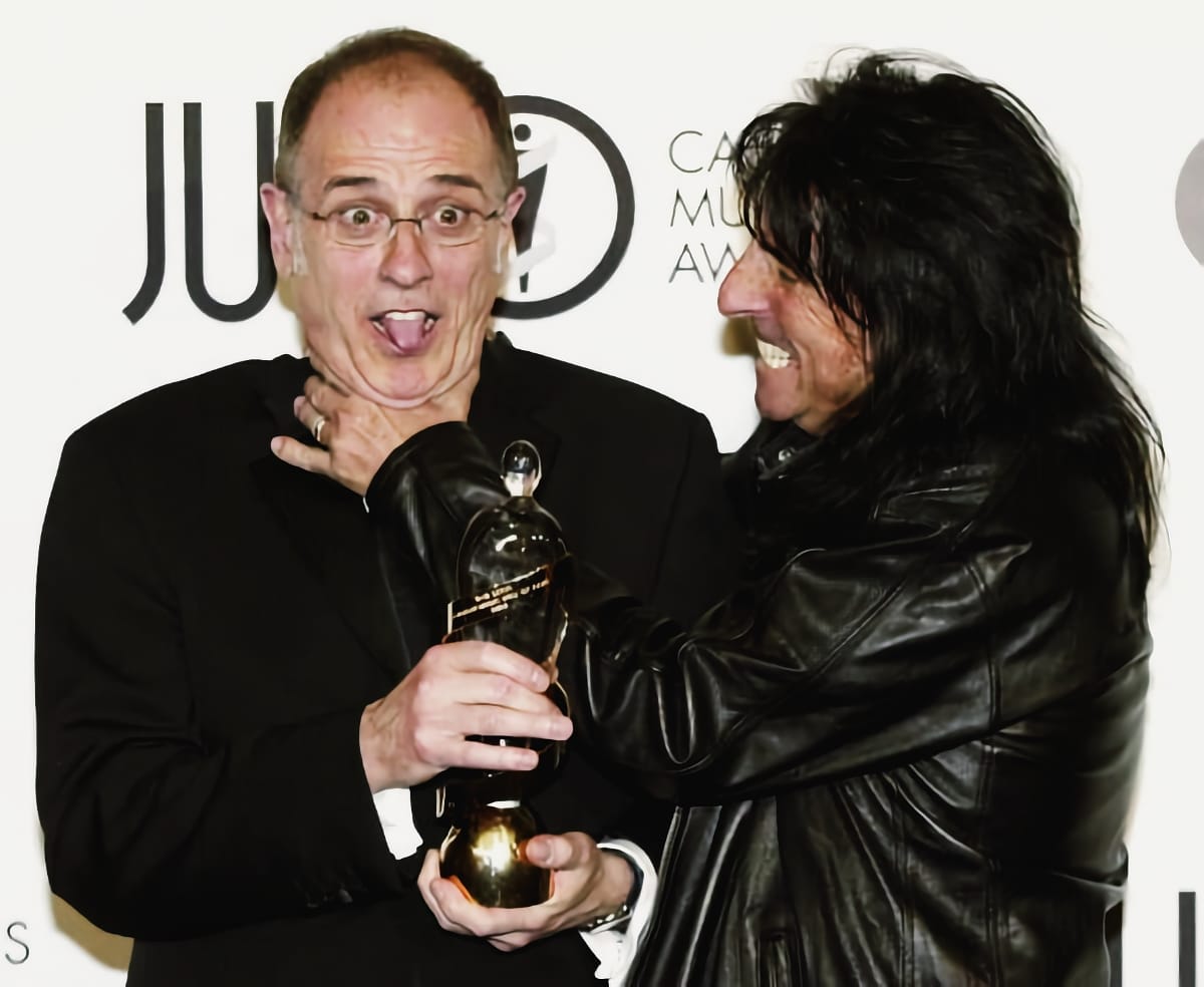 Bob Ezrin, eyes wide and tongue out, holds a gold Juno Award while Alice Cooper grins and puts a hand around his throat, the Juno Awards backdrop behind them.