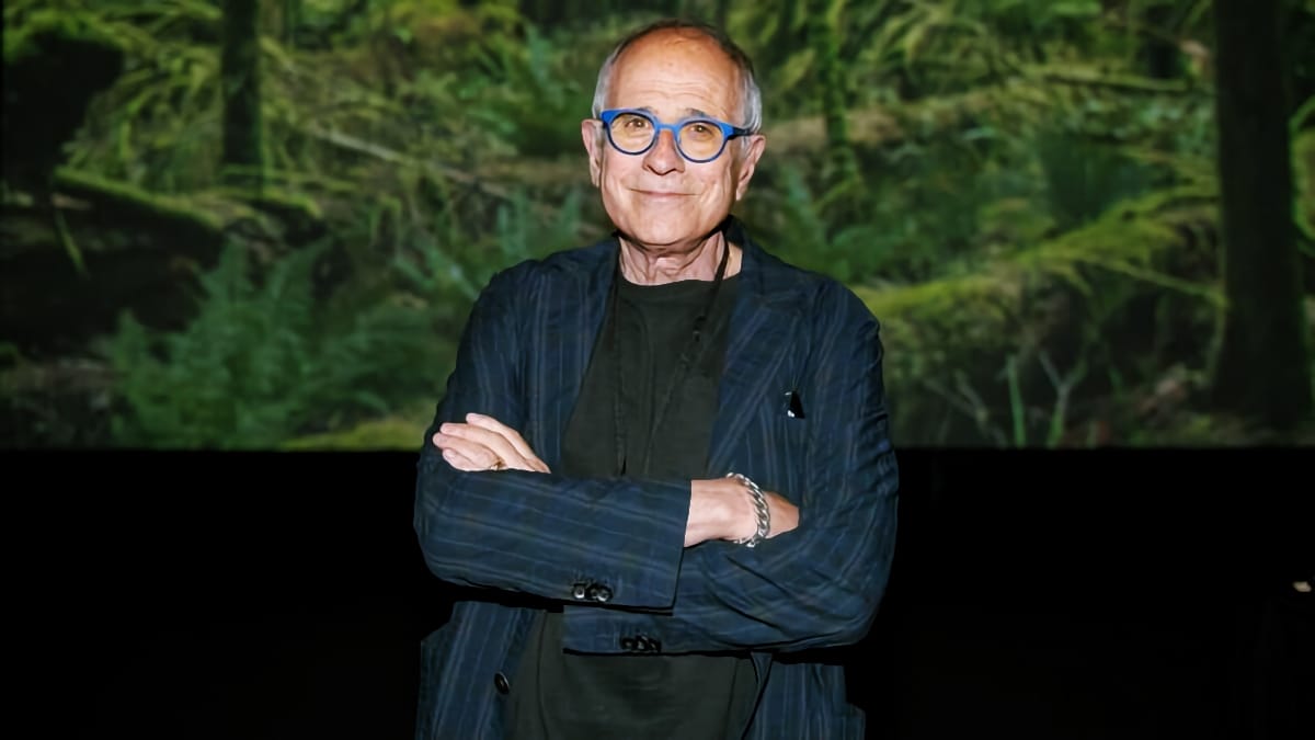 Bob Ezrin stands with his arms folded in blue-framed glasses and a dark pinstriped jacket, a projected image of mossy forest filling the screen behind him.