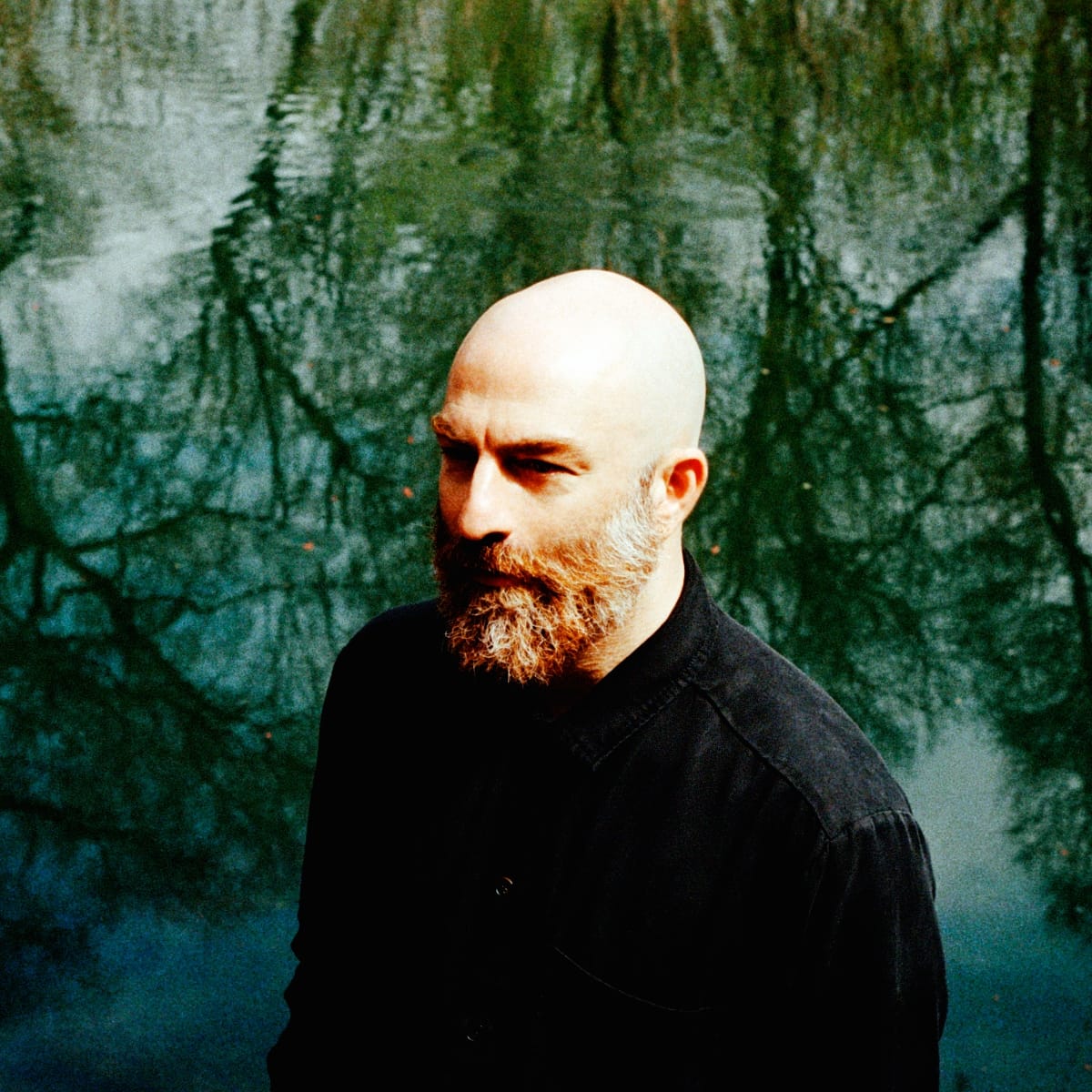Deniz Cuylan in profile, looking off to his left, against dark green water with bare branches reflected in it.