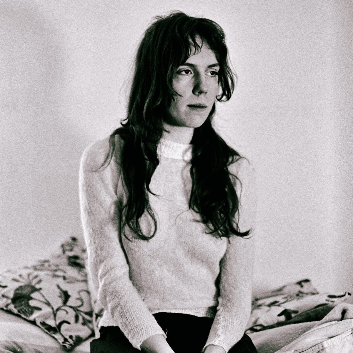 Elanor Moss sits on a bed of floral-embroidered cushions in a soft pale sweater and dark trousers, long hair loose, looking past the camera. Grainy black-and-white portrait.