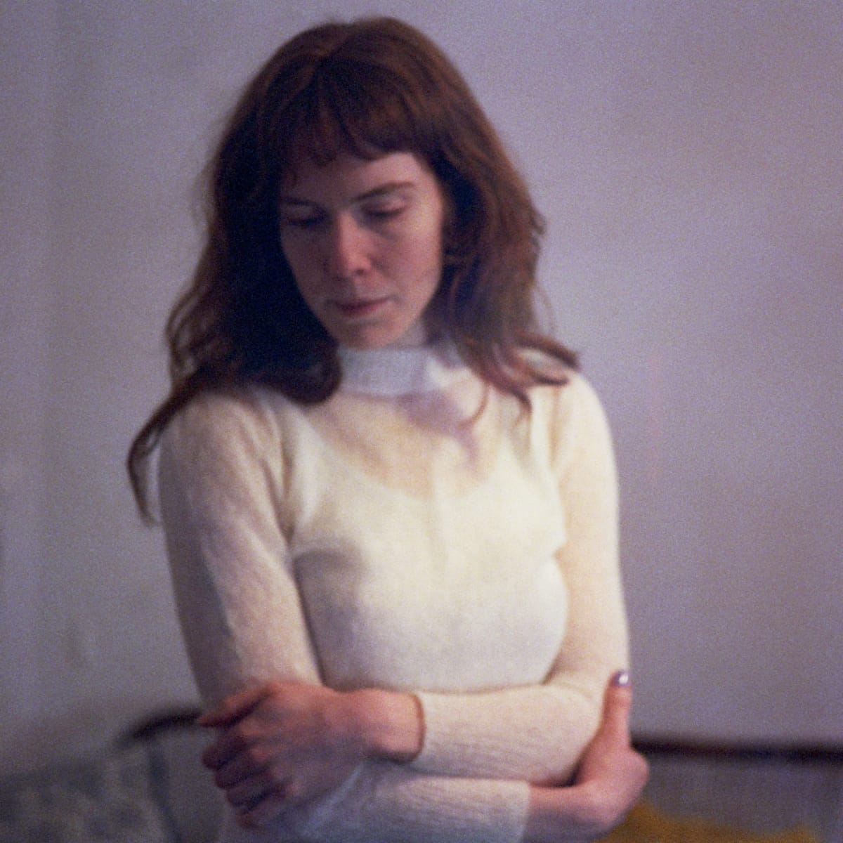 Elanor Moss stands with her arms folded against a pale wall, head lowered, in a cream mohair sweater. Soft focus and warm light blur the edges of the frame.
