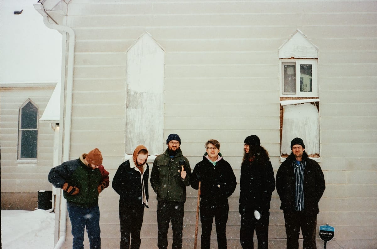 Fling ii, six members in winter coats and knit caps, standing in a row against the white clapboard wall of a church as snow falls around them. One holds a wooden walking stick.