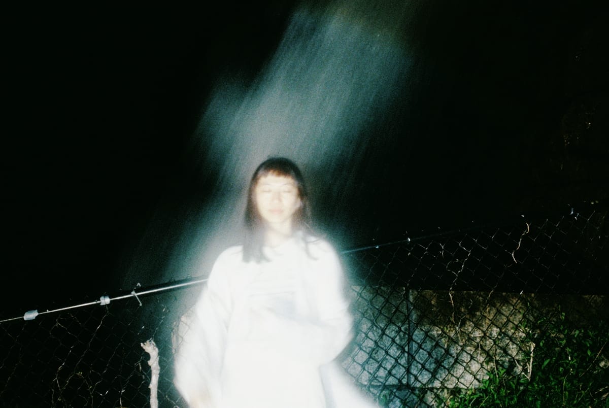 Frances Chang stands at night before a chain-link fence in a white top, eyes closed, a streak of light falling diagonally across the frame and blurring her outline.