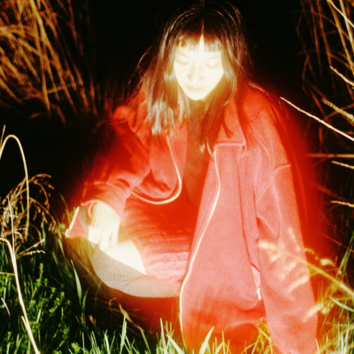 Frances Chang crouches in tall grass at night in a red jacket, head lowered toward a light held in her hands, a red flare washing across the frame.