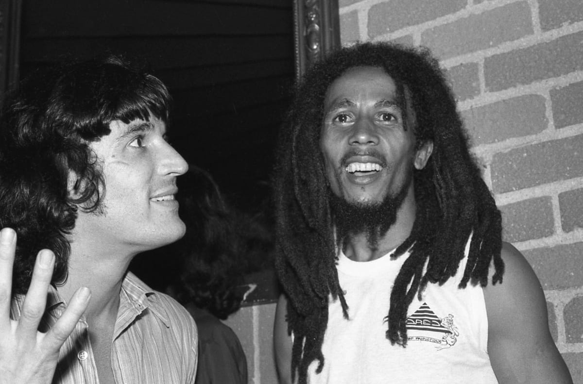 Rick Danko and Bob Marley stand against a brick wall, Danko turned toward him mid-sentence with one hand raised, Marley laughing and looking off past the camera.