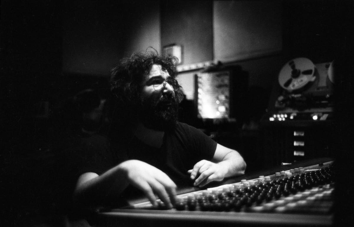 Jerry Garcia sits at a recording console in a dark studio, one hand resting on the faders, a reel-to-reel tape machine behind him.
