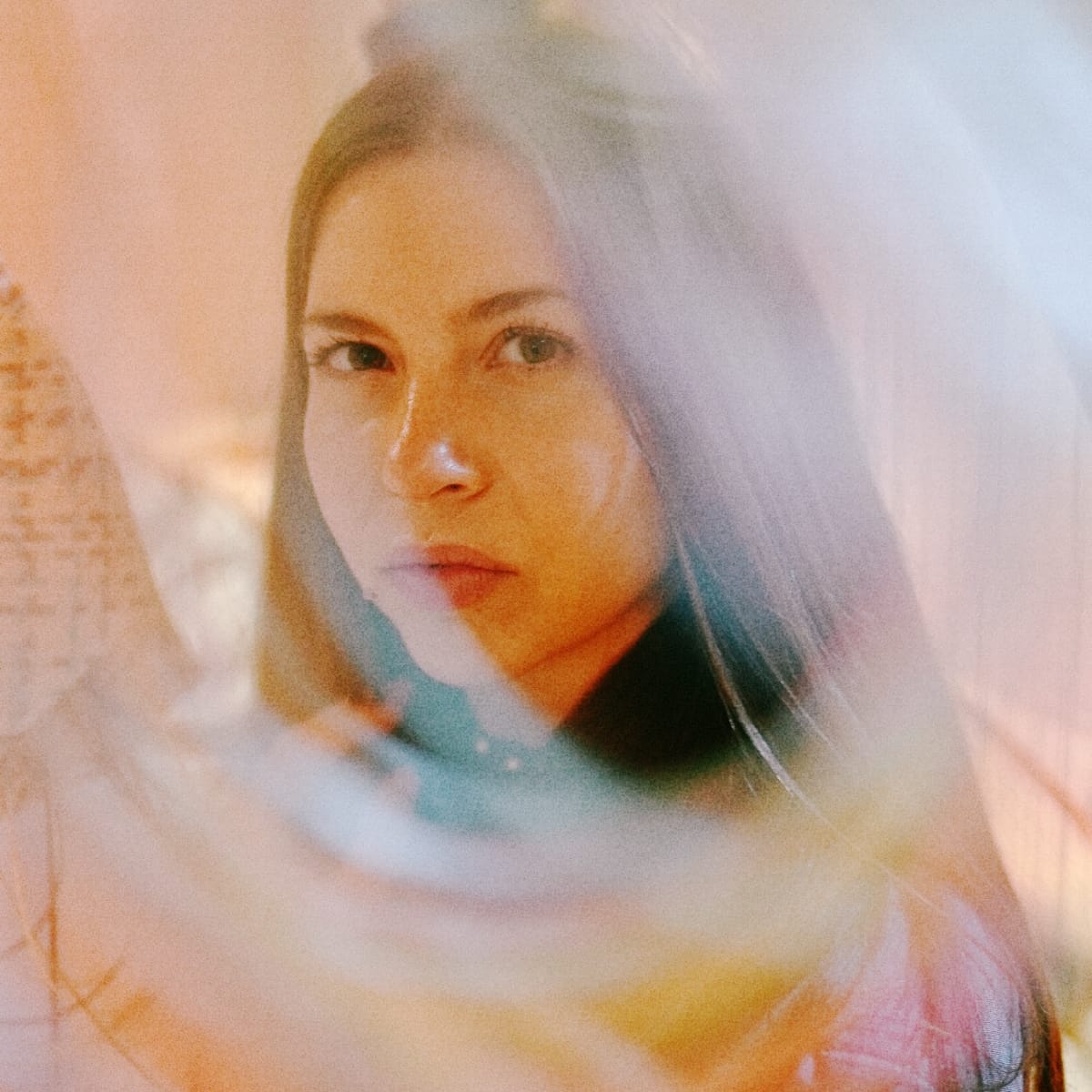 Lara Somogyi looks straight at the camera through a soft blur of translucent pastel fabric, her face framed by long hair in warm peach light.