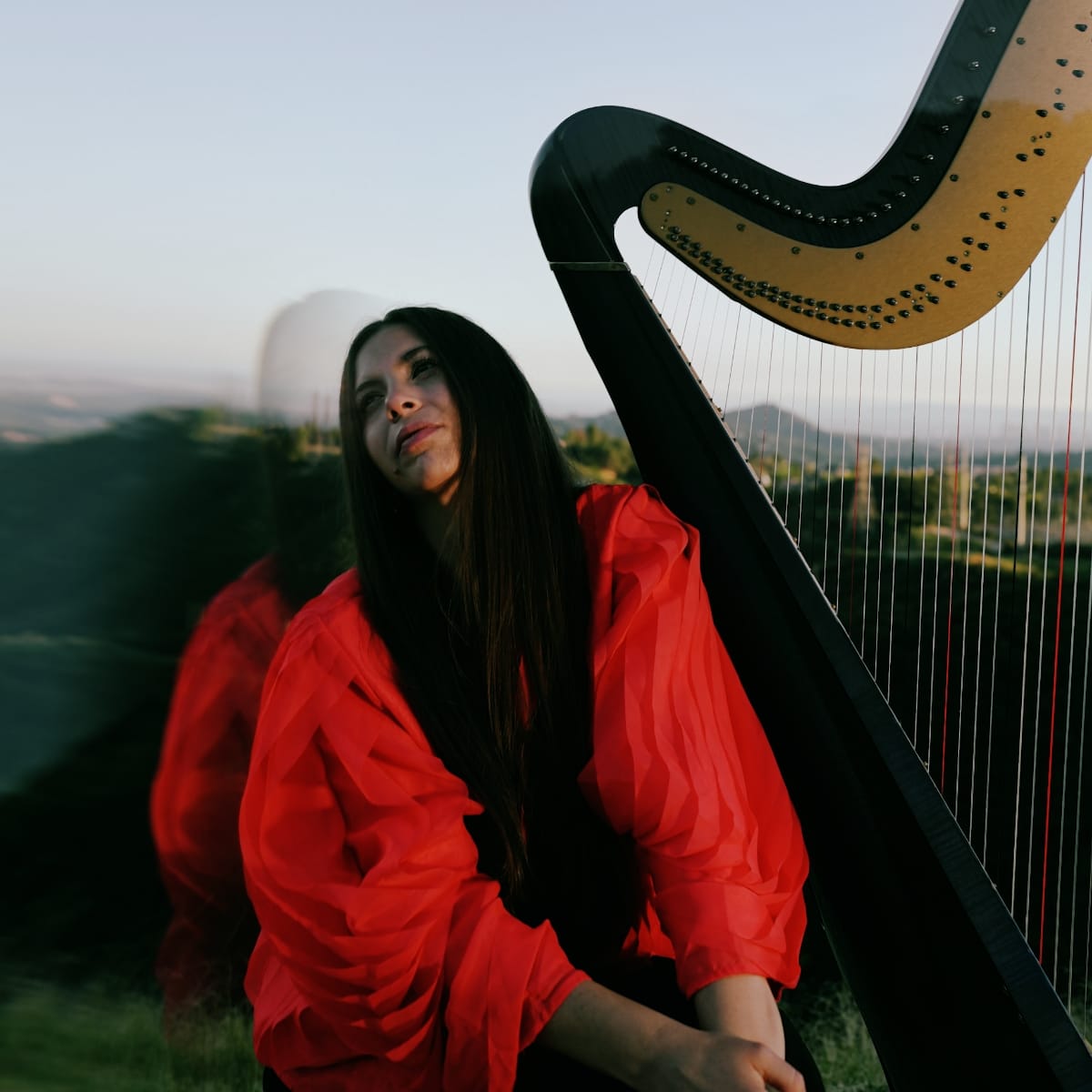 Lara Somogyi in a flowing red blouse leans against the column of a black-and-gold concert harp on a hillside at dusk, a faint double exposure echoing her figure.