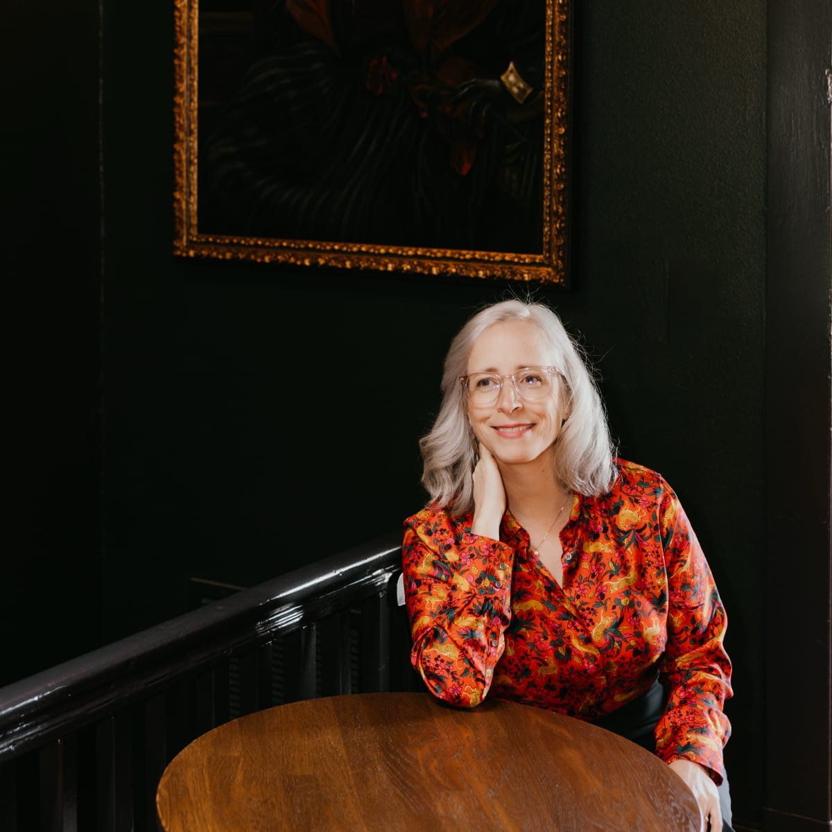 Laura Veirs sits at a round wooden table, chin propped on one hand, smiling, in a red floral shirt against a dark green wall hung with a gilt-framed painting.