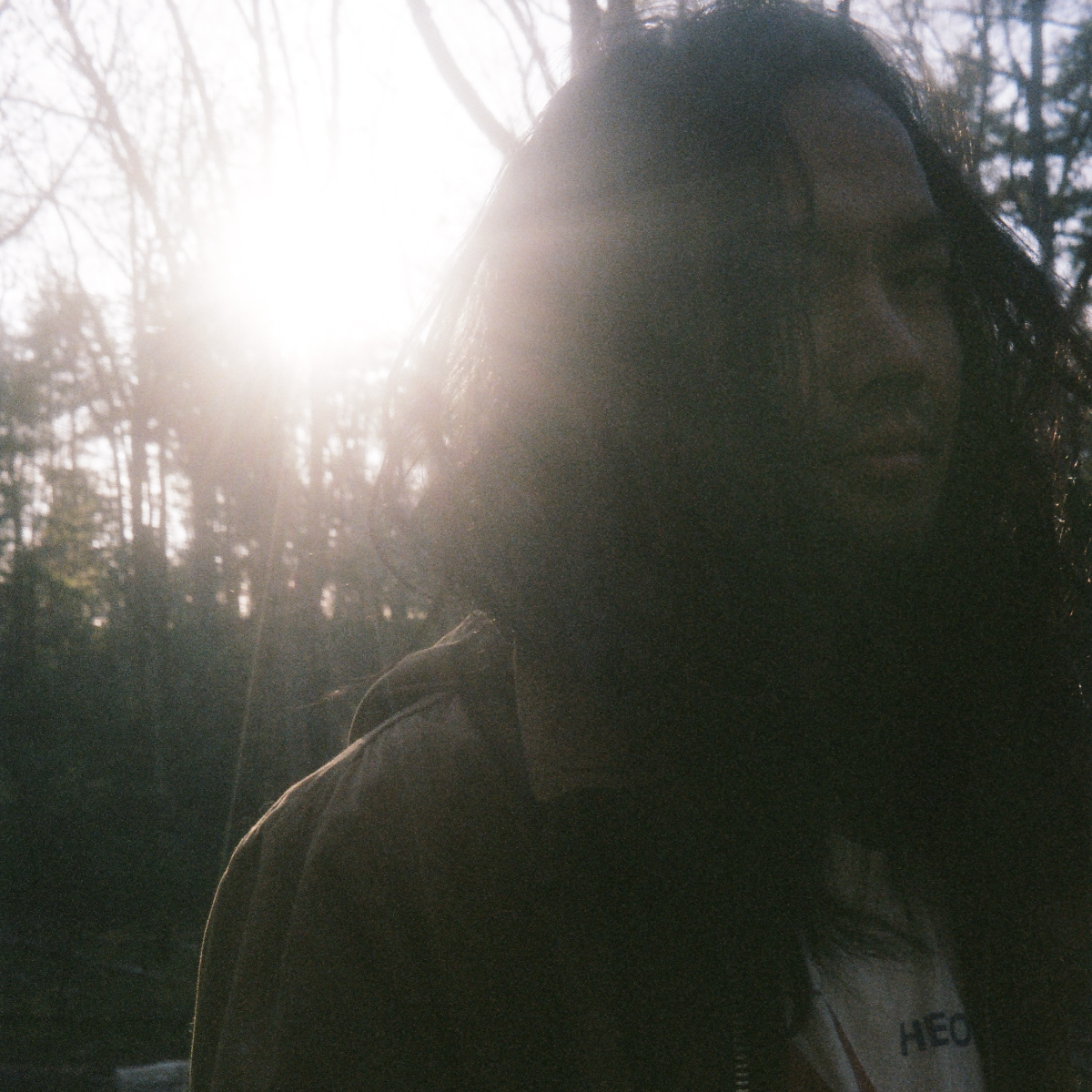 Christian Li looks off to one side in bare winter woods, long dark hair falling across his face, low sun flaring behind him and leaving his features in shadow.