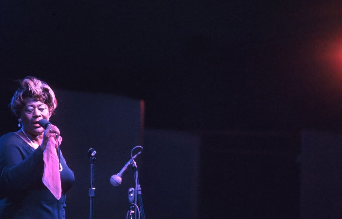 Ella Fitzgerald sings into a handheld mic at the Newport Jazz Festival, lit deep magenta on a dark stage, a scarf gathered in one hand.