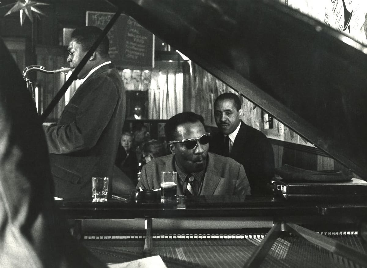 Thelonious Monk sits at an open grand in dark sunglasses at The 5 Spot in 1957, John Coltrane playing tenor behind him and drummer Shadow Wilson leaning over his shoulder.