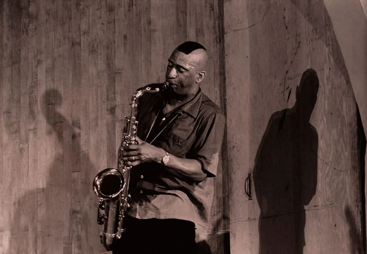 Sonny Rollins plays tenor saxophone against a weathered wood wall at the Museum of Modern Art, his shadow and a second figure's thrown large beside him.