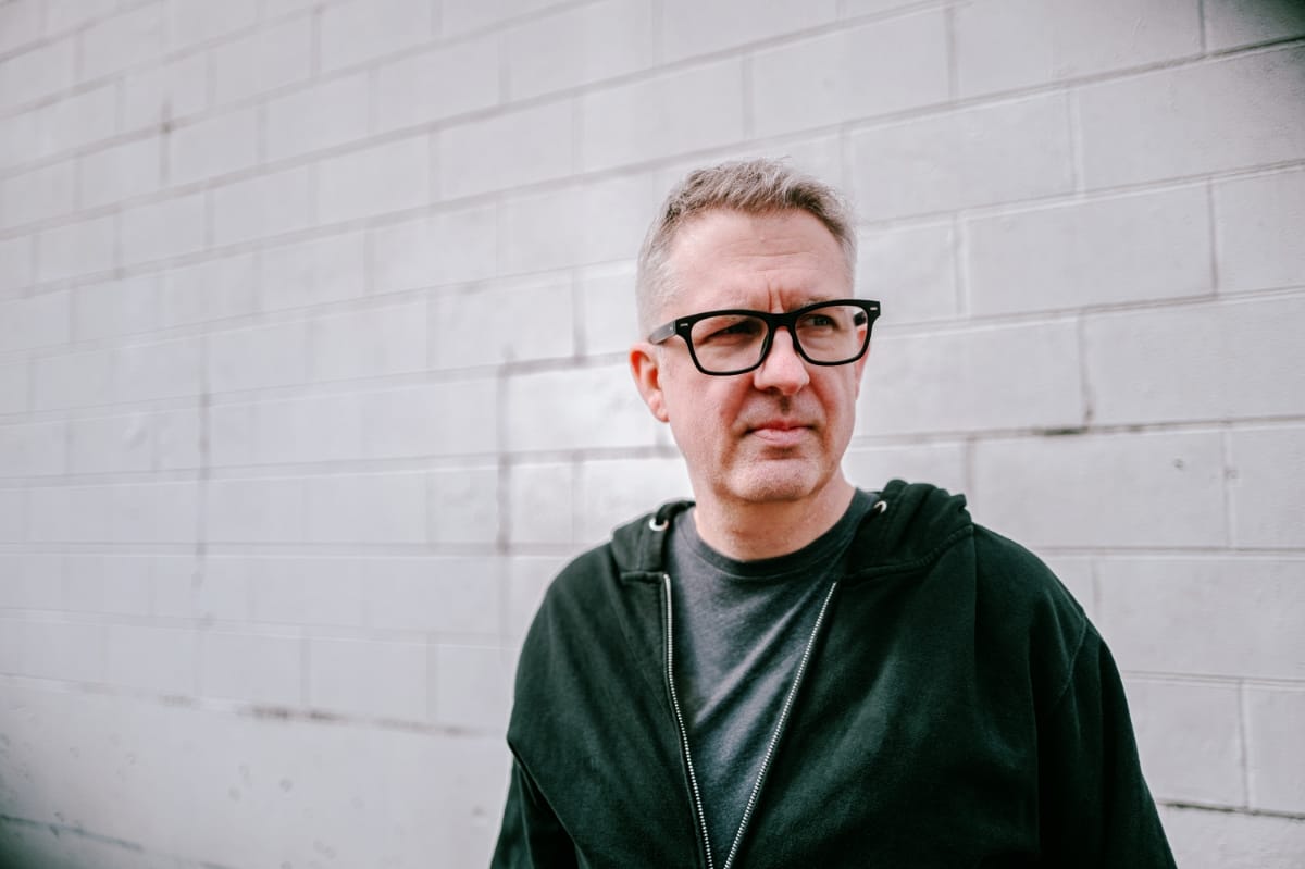Jeff McIlwain, aka Lusine, in a dark hooded jacket and black rectangular glasses, standing before a white-painted cinder block wall and looking past the camera.