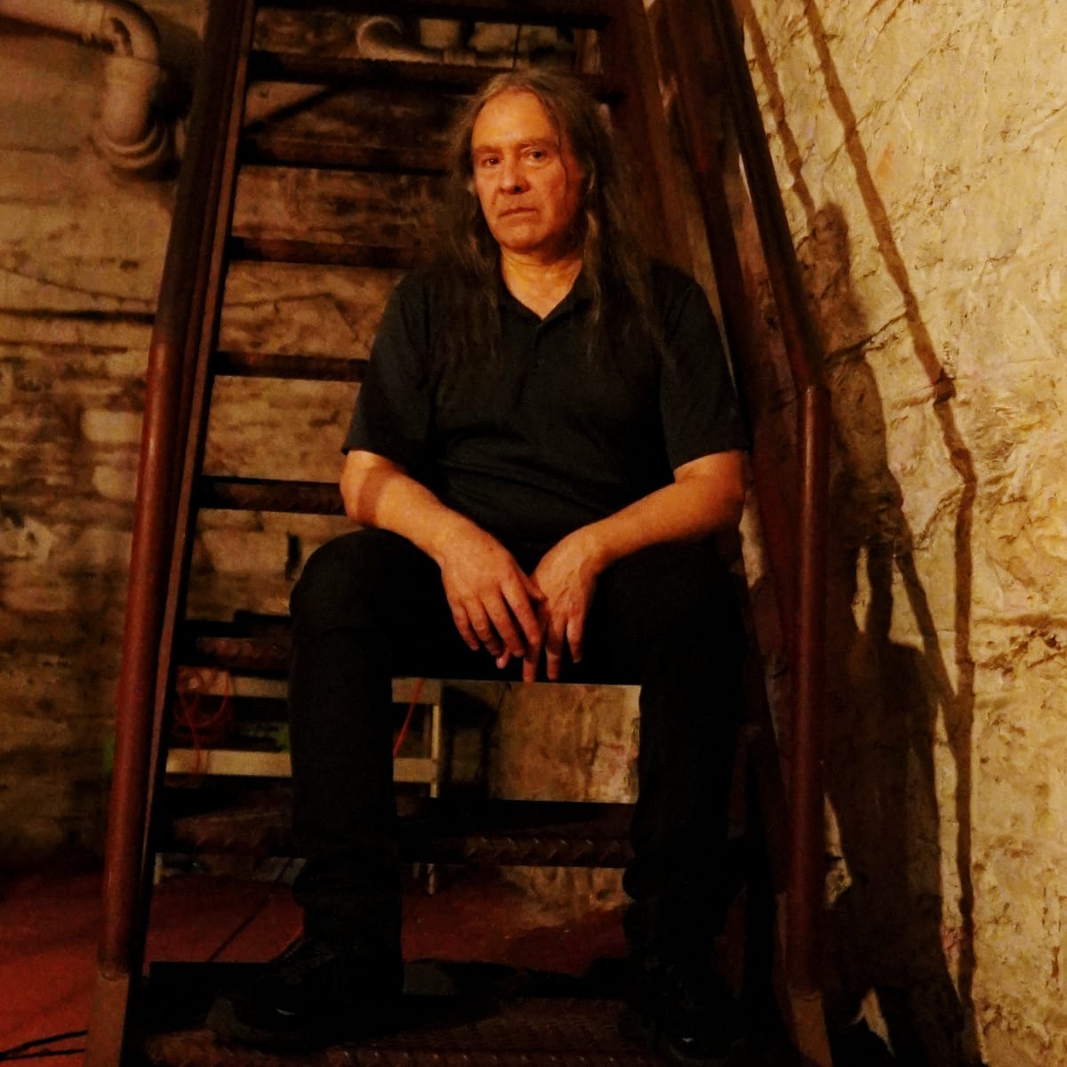 Martin Bisi sits on a steep, narrow staircase in a dim basement, hands clasped between his knees, warm light picking out old brick, plaster, and overhead pipes.