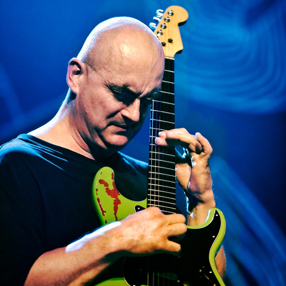 Olaf Rupp plays a lime-green Stratocaster held almost upright, head bowed and eyes lowered, lit against a swirl of deep blue stage ligh